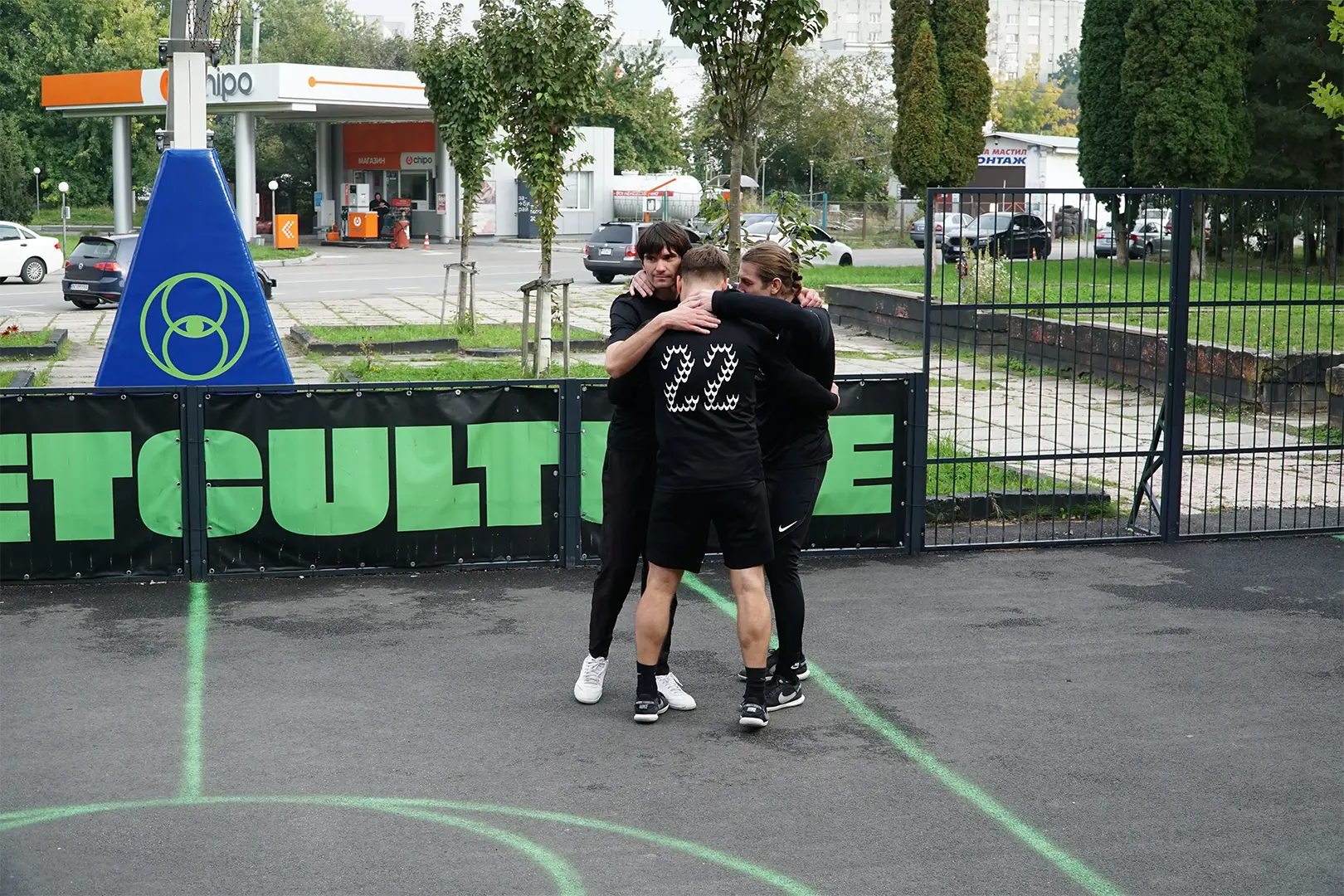 Trzej członkowie ekipy Warsaw FC w ciasnym, grupowym uścisku na środku betonowego boiska we Lwowie. Zdjęcie symbolizuje jedność, wsparcie i braterską więź zespołu podczas turnieju.