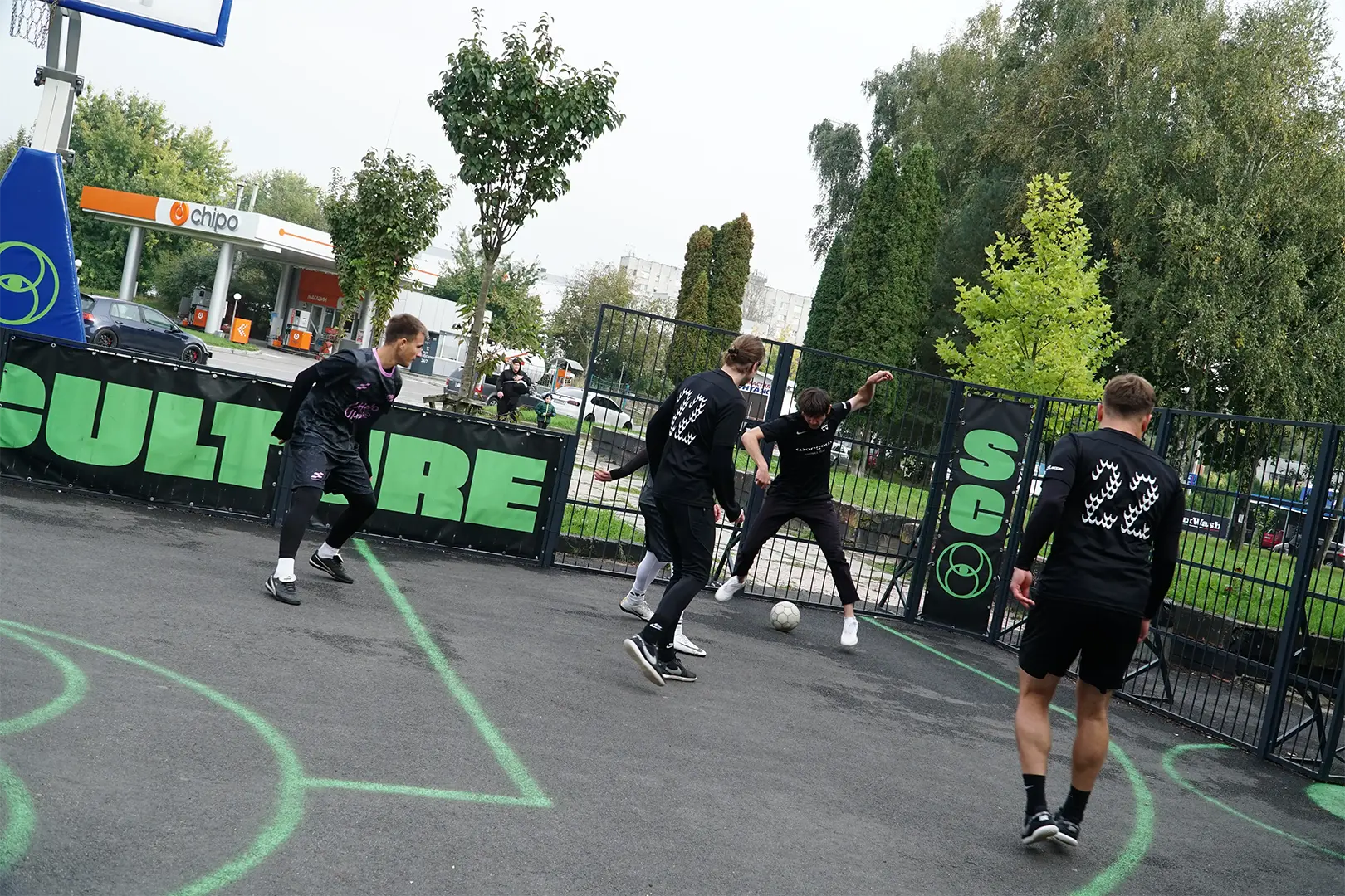 Dynamiczne ujęcie meczu street footballu na mokrym betonowym boisku we Lwowie. Zawodnicy Warsaw FC walczą o piłkę wewnątrz klatki, w tle widoczny baner z napisem CULTURE i stacja benzynowa.