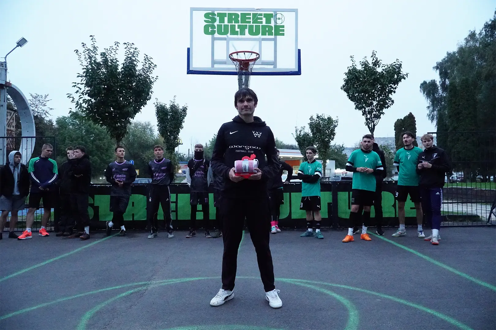 Michał Rycaj, zawodnik Warsaw FC, stoi na betonowym boisku we Lwowie, trzymając w rękach różową statuetkę MVP turnieju street footballu. W tle widoczni są inni uczestnicy turnieju oraz kosz do koszykówki z napisem STREET CULTURE.