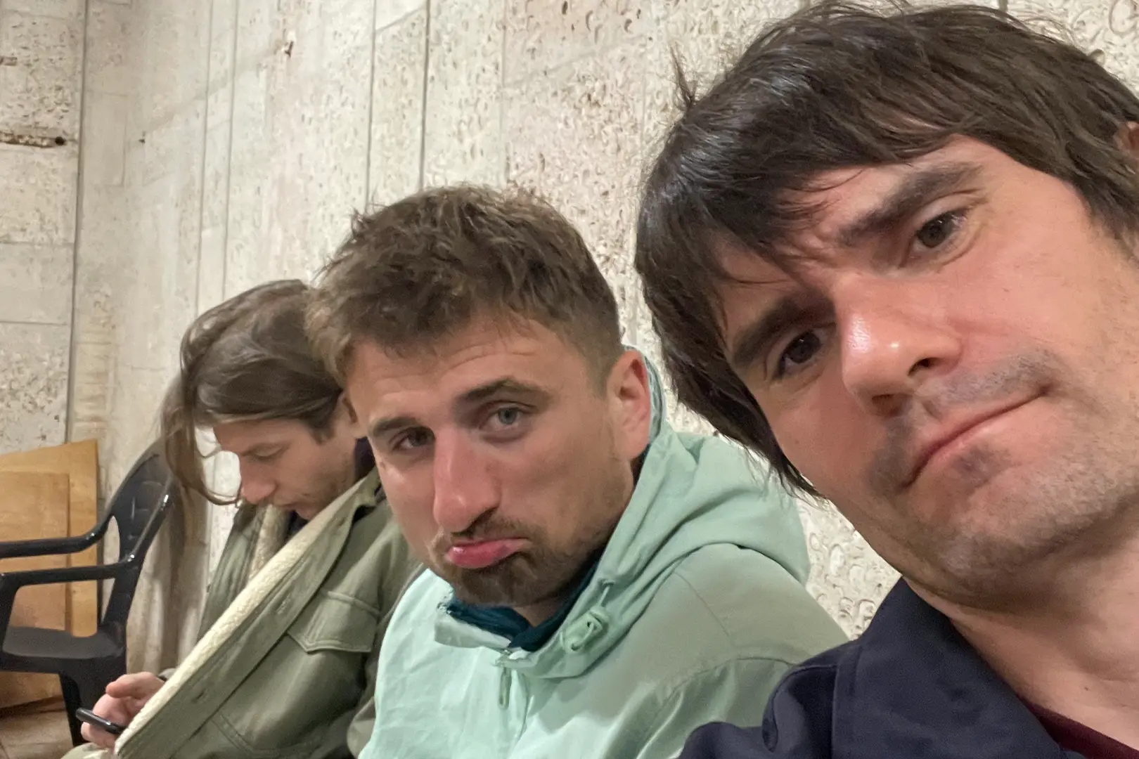 Selfie zrobione w surowym, betonowym schronie we Lwowie podczas nocnego alarmu rakietowego. Od lewej siedzą członkowie ekipy Warsaw FC: Damian Gawrych (patrzy w dół), Karol (zrezygnowany wyraz twarzy) i Michał Rycaj (autor zdjęcia). Ich twarze zdradzają zmęczenie i napięcie po kilku godzinach spędzonych pod ziemią.