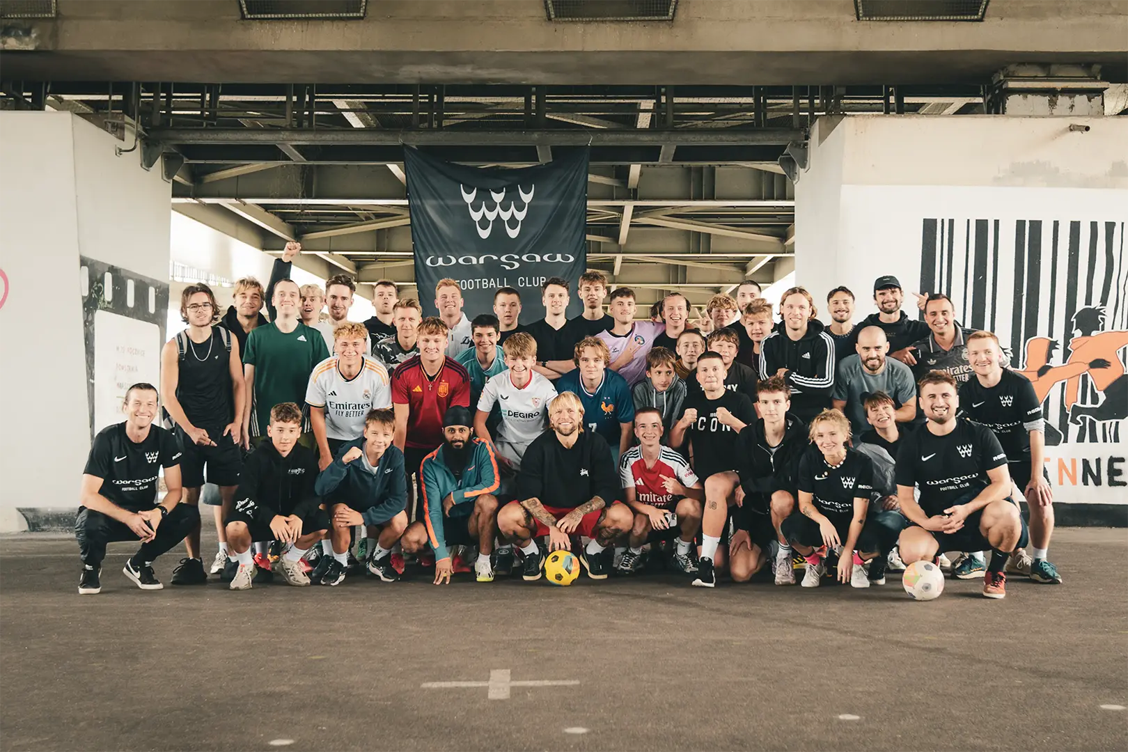 Zdjęcie grupowe społeczności street footballowej Warsaw FC na betonowym boisku pod mostem łazienkowskim w Warszawie. W centralnym punkcie siedzi piłkarz reprezentacji Polski, Tymoteusz Puchacz, trzymający piłkę. W tle widoczny baner Warsaw Football Club.
